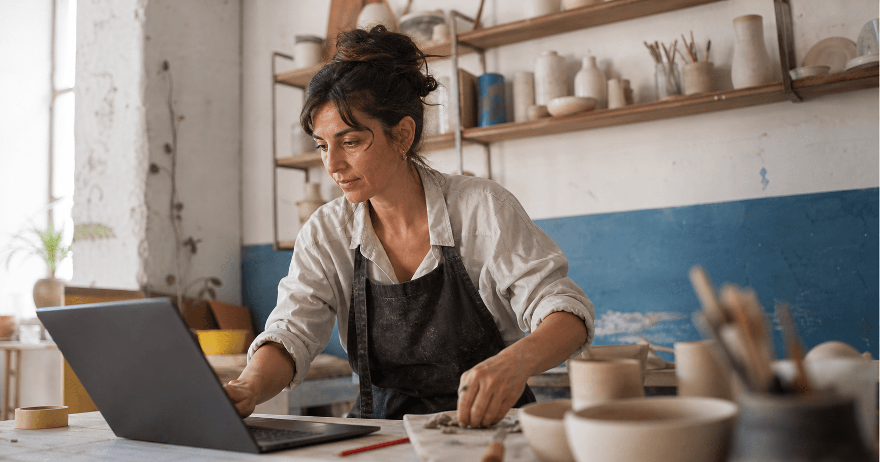 Artesana utilizando un portátil mientras trabaja en su taller de cerámica